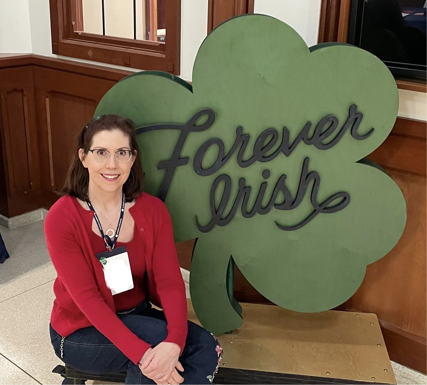 a woman with brown hair is sitting on a bench in front of a four-leaf clover sign. the woman is waring glasses, lanyard, red long sleeve shirt, and black pants.
