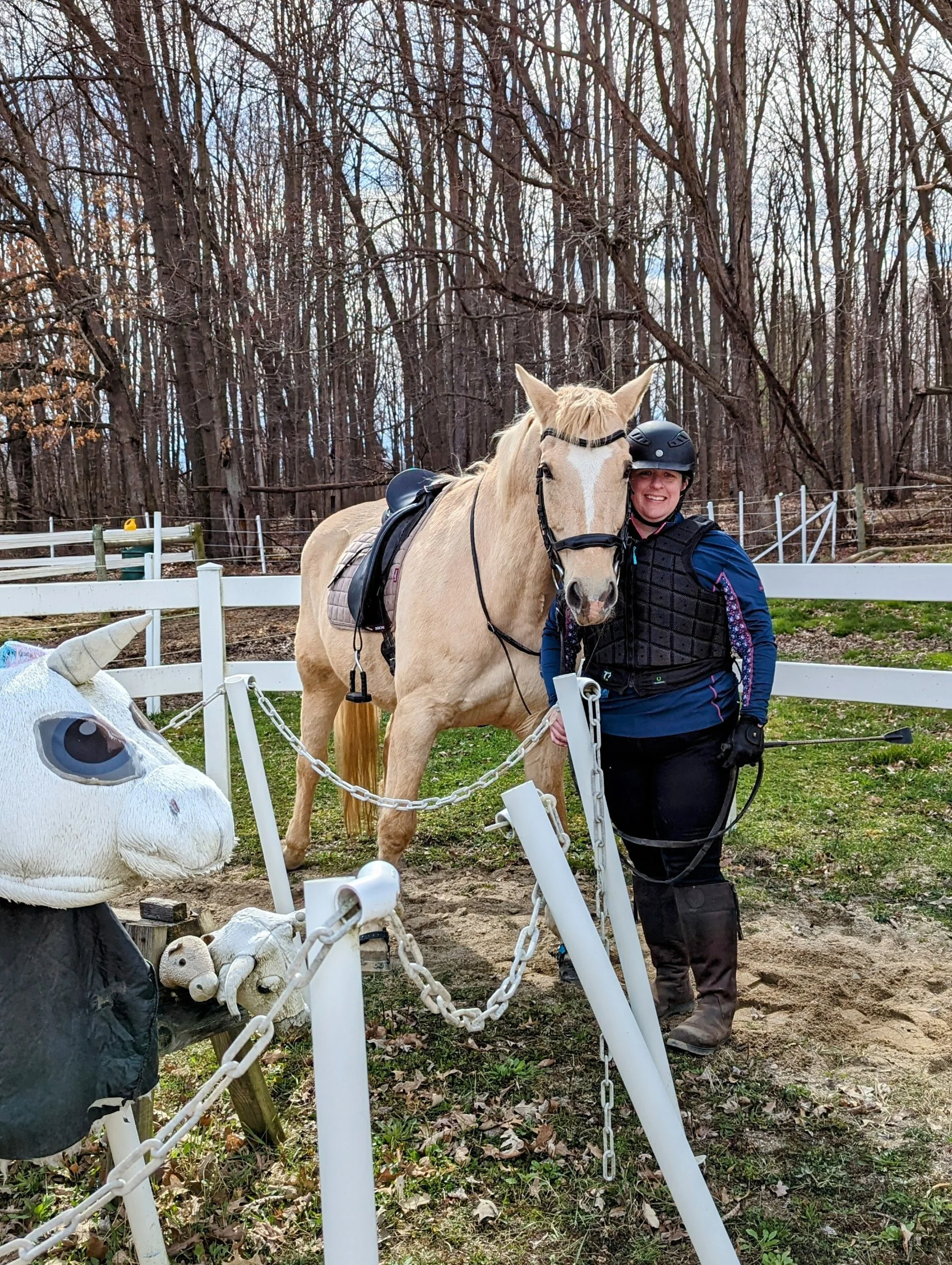 a woman stands outside in front of a white fence, holding a tan horse and smiling. the woman is wearing a black helmet, black long sleeved shirt, black pants, and boots.