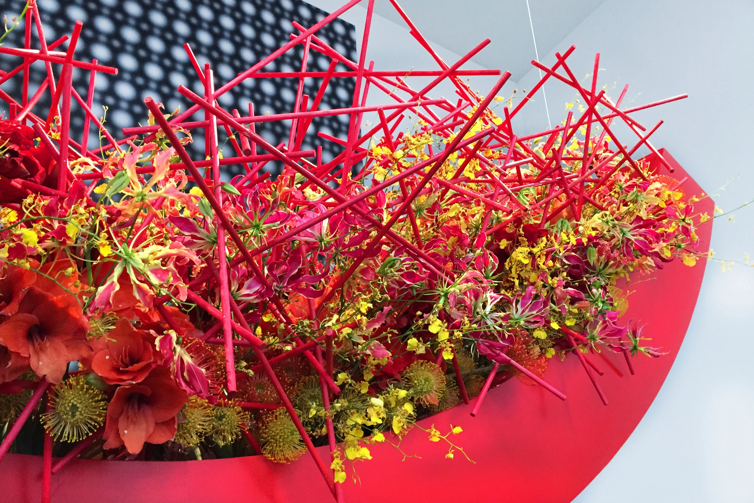 A colorful floral arrangement with red, yellow, and pink flowers in a red wrapping, accented by a red decorative structure, set against a background with a patterned dark and light surface and a light blue sky.