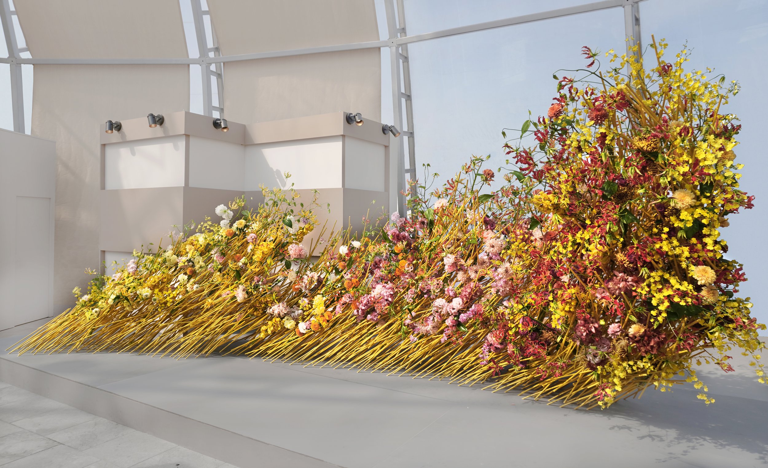A modern indoor floral art installation with yellow, pink, and red flowers arranged on a slanted golden structure, set against a white wall and a glass window ceiling.