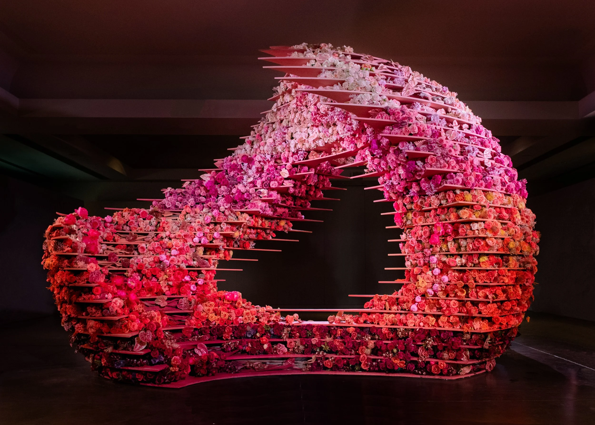 An artistic floral installation made of horizontal pink shelves filled with pink and white flowers, creating a curved architectural structure in a gallery or indoor space.