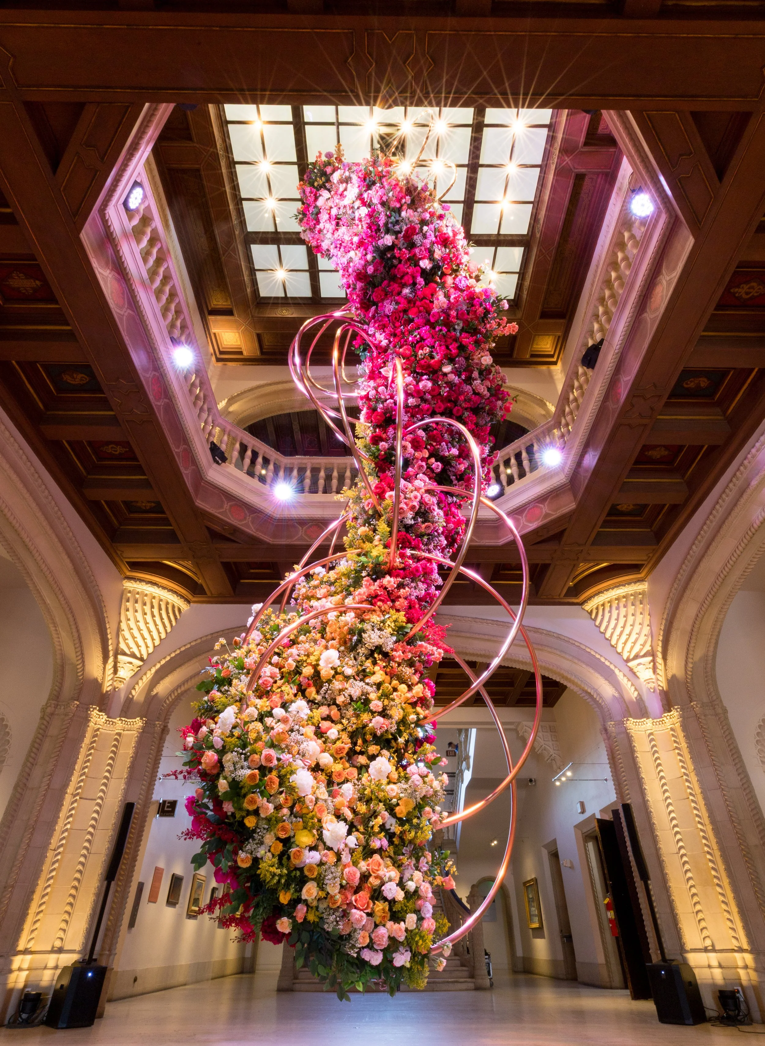 A tall floral arrangement featuring pink, white, orange, and yellow flowers hangs from the ceiling in a grand lobby with ornate architectural details and a skylight.