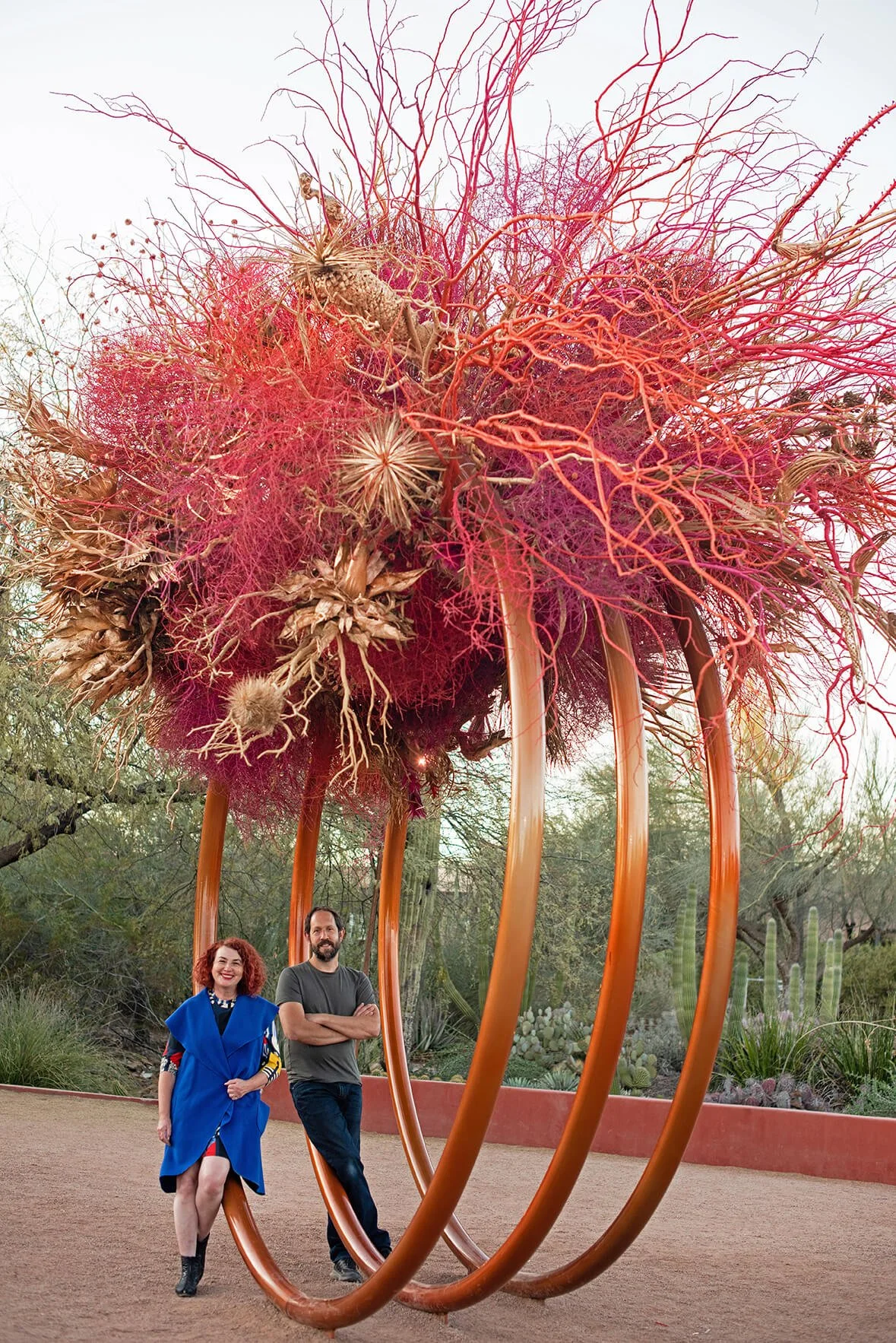 Two people, a woman with curly red hair wearing a colorful dress and a blue scarf and a man with a beard wearing a gray t-shirt, standing in front of a large outdoor sculpture resembling a bouquet of pink and red thistles. The sculpture is supported by curved orange metal arches and is set in a desert landscape with cacti and desert plants.