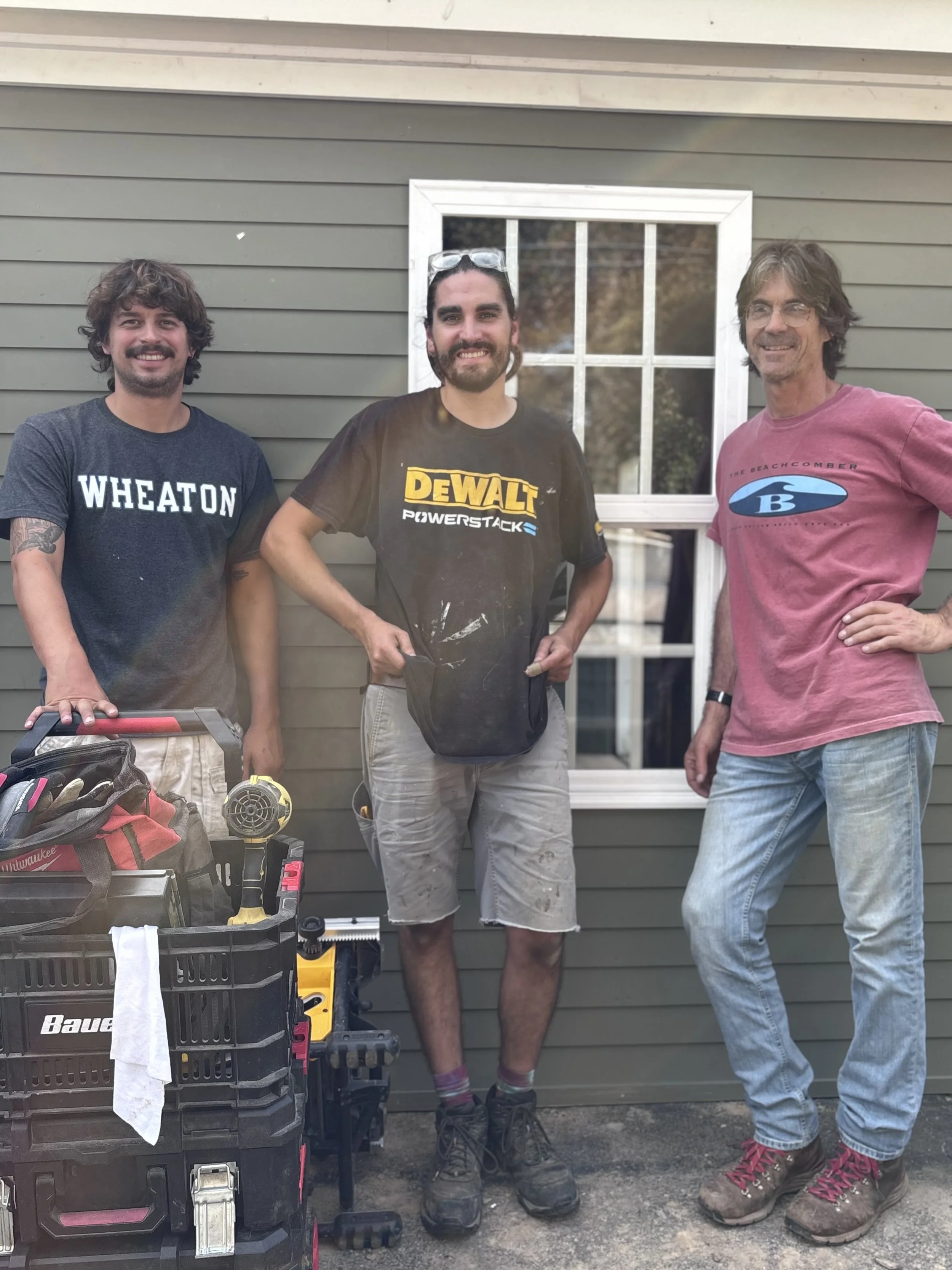 Three crew members standing beside tools outside a house.