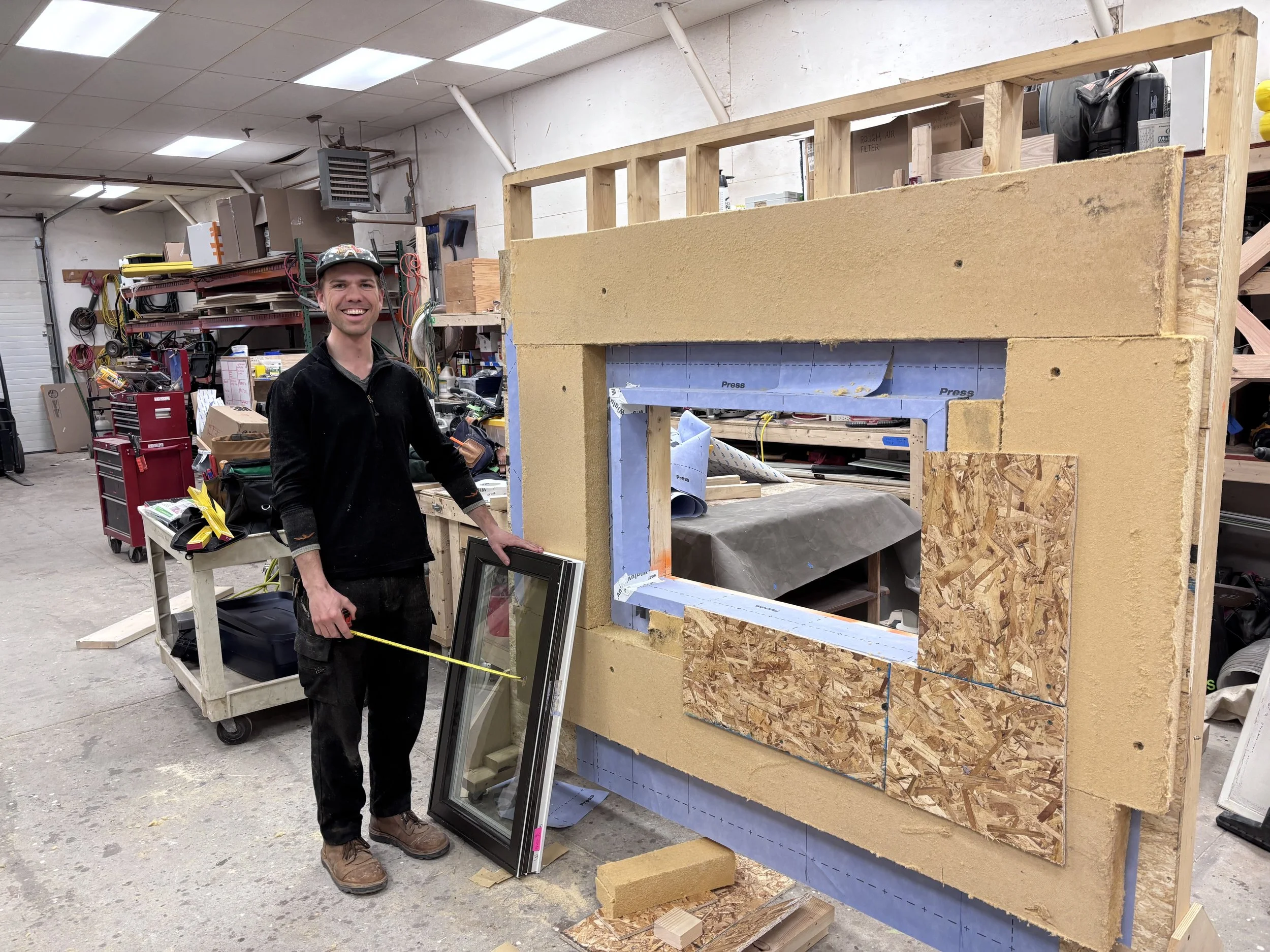 Carpenter beside a wall mock-up showing insulation and a window opening in a workshop.