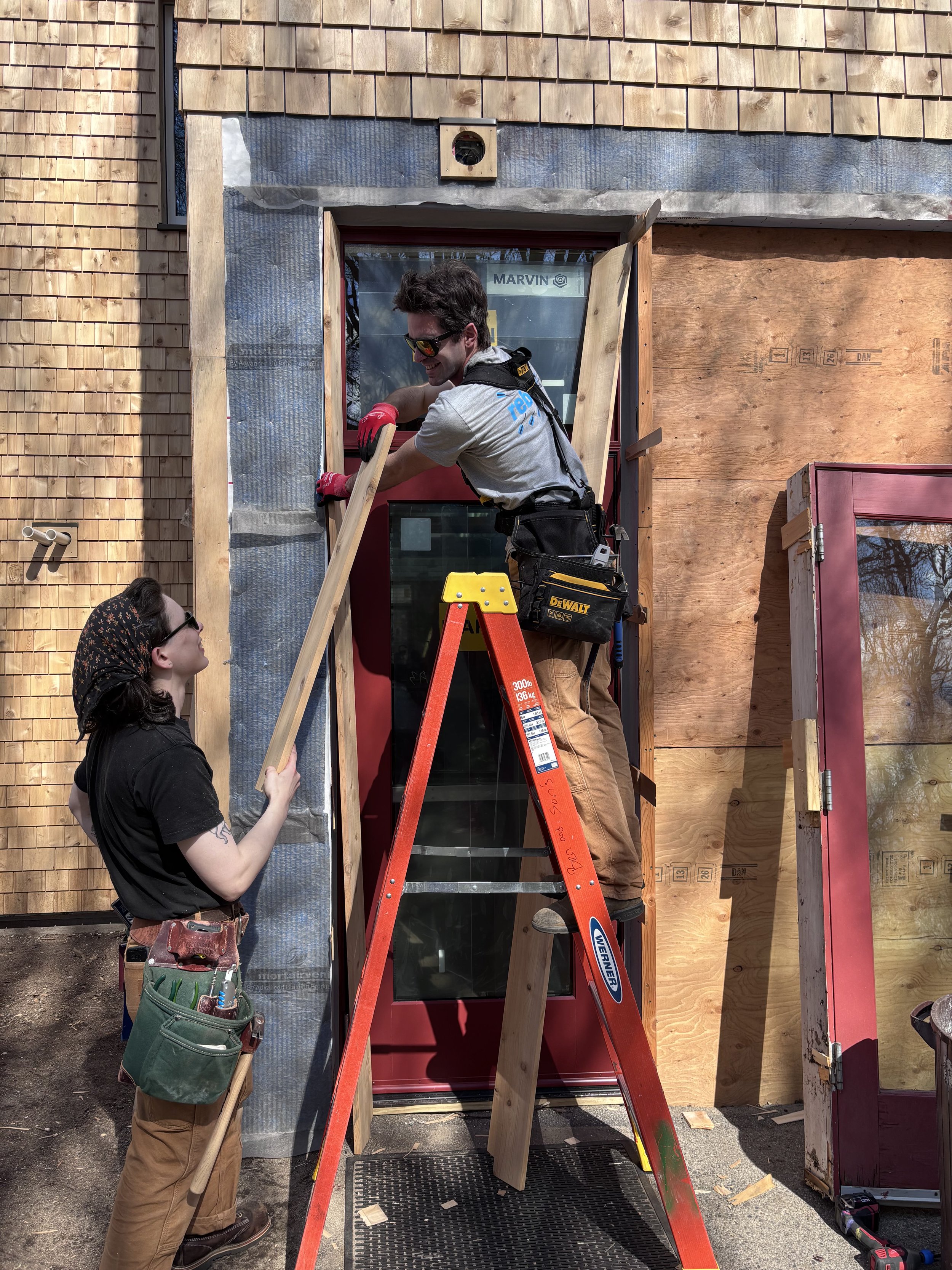Two carpenters working at a window opening, with a ladder and exposed exterior wall layers.