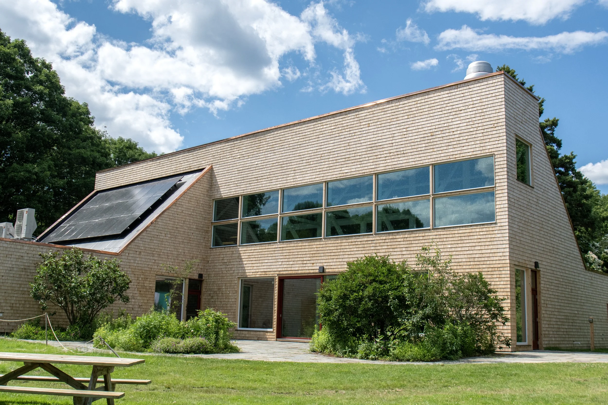 Shingled building with large windows, solar panels, and a picnic table on the lawn.
