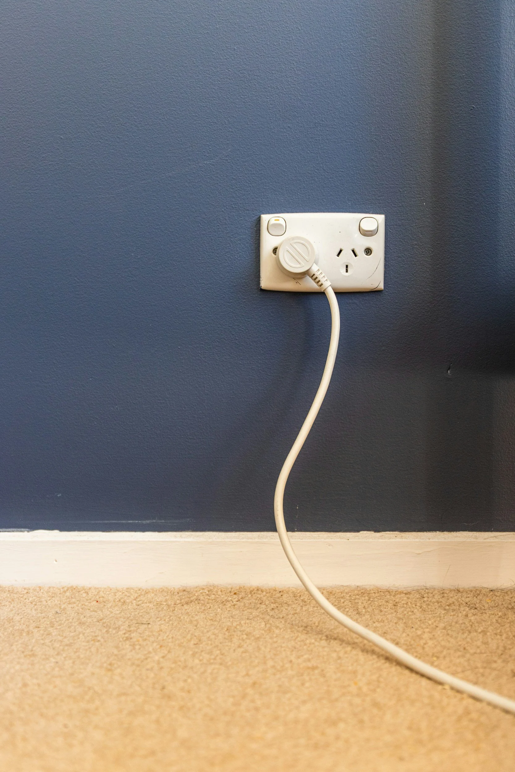 Electric outlet with a plugged-in white power cord on a blue wall, near beige carpet.