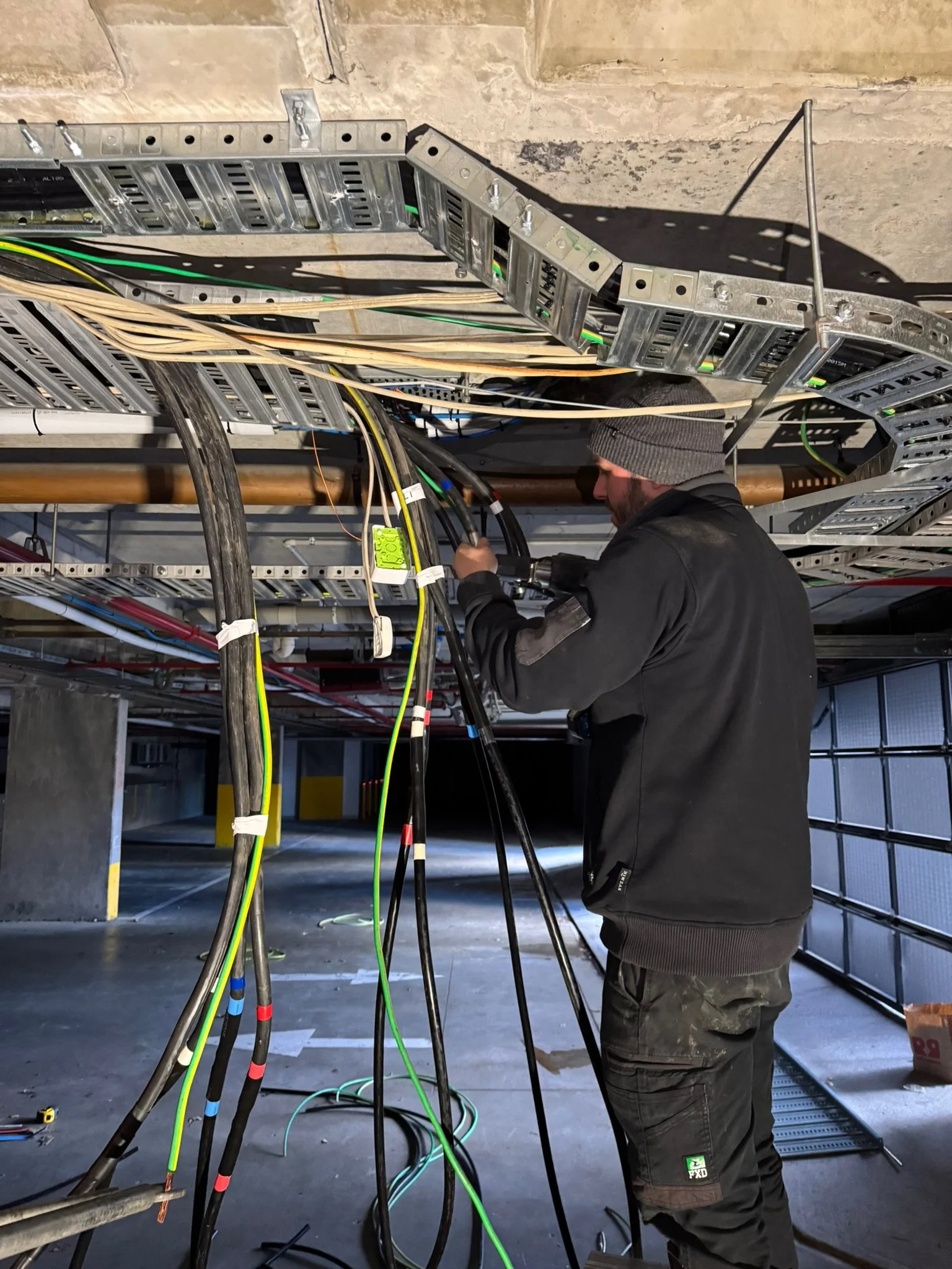 A person with a gray beanie and black jacket working on electrical wires at a construction site with metal framing and wiring overhead, in a partially finished building.