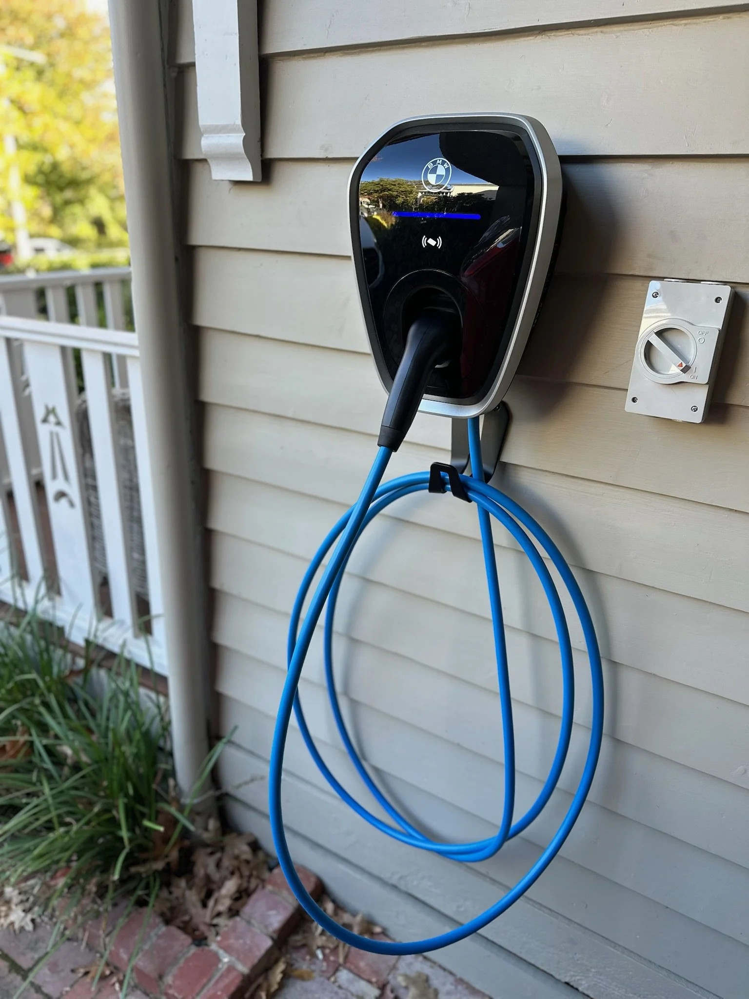 An electric car charging station mounted on the exterior wall of a house, with an electric BMW car brand logo visible and a blue charging cable hanging.