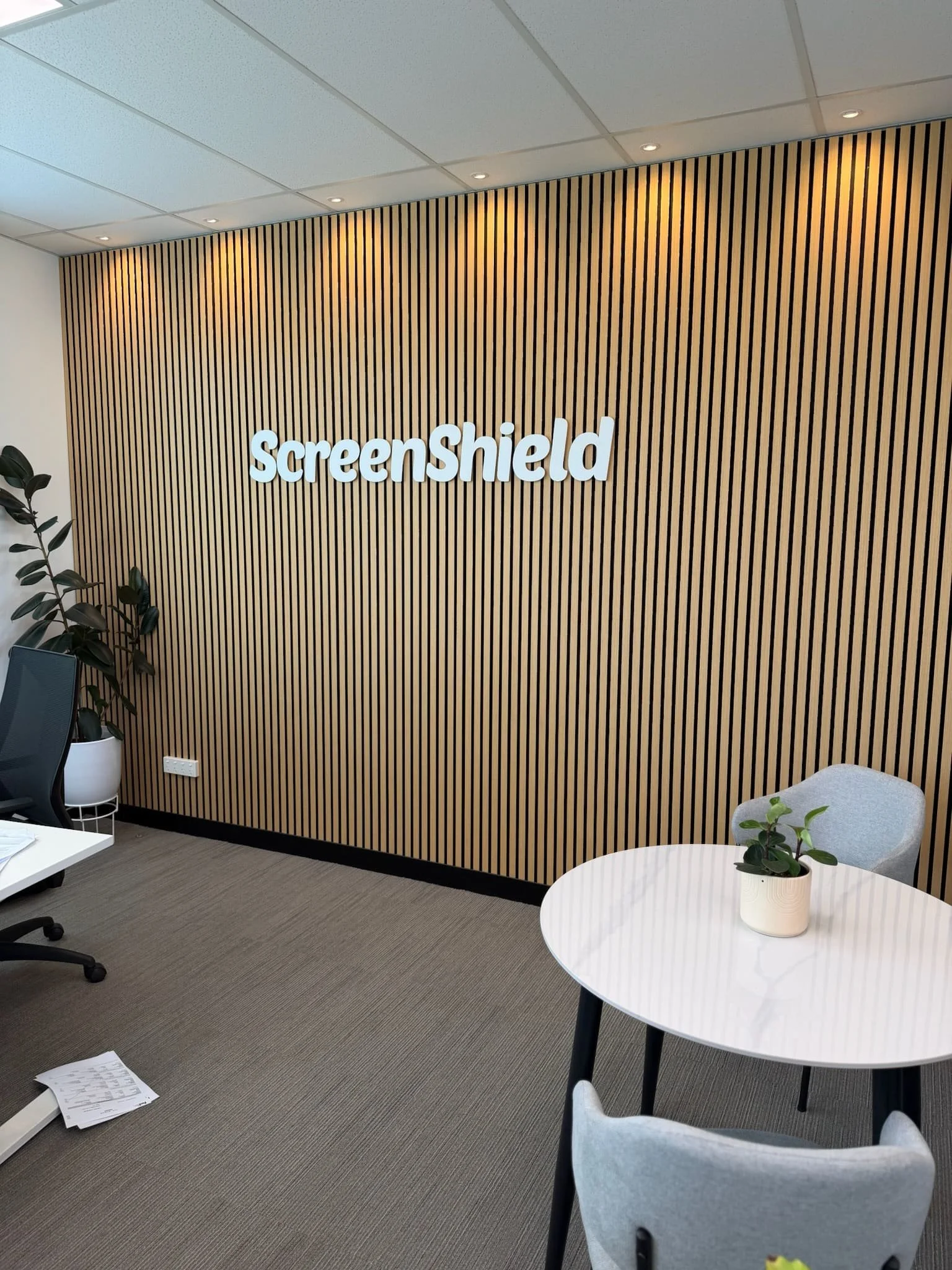 Office space with a wooden slat accent wall displaying the company name 'ScreenShield' in white letters. There are white tables with small potted plants, chairs, and office supplies, with a ceiling featuring spotlights.