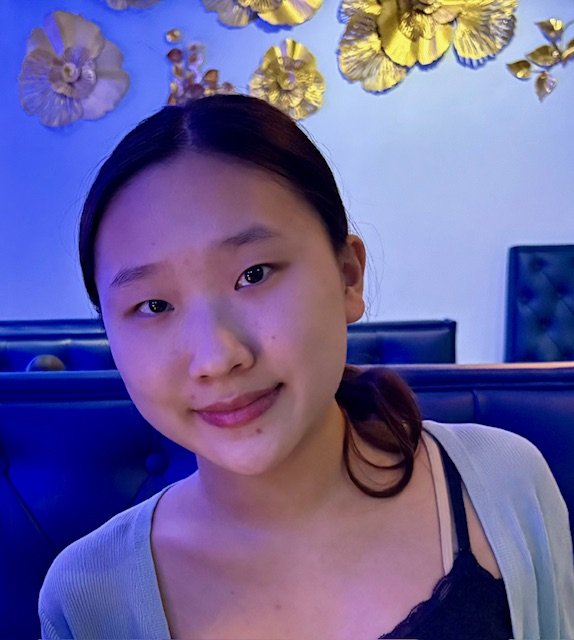 A girl with dark hair pulled back smiles, while the wall behind her has gold flowers on it