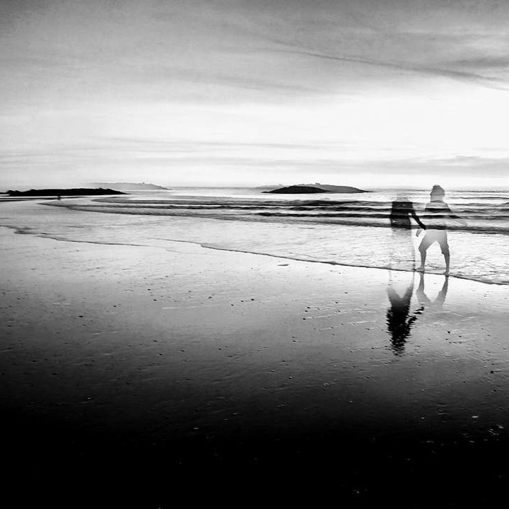  The image is black and white and mainly a landscape photograph of the beach with calm waves. The image shows the silhouette / faint outline of Fin and they are stood in the water with bare feet. 