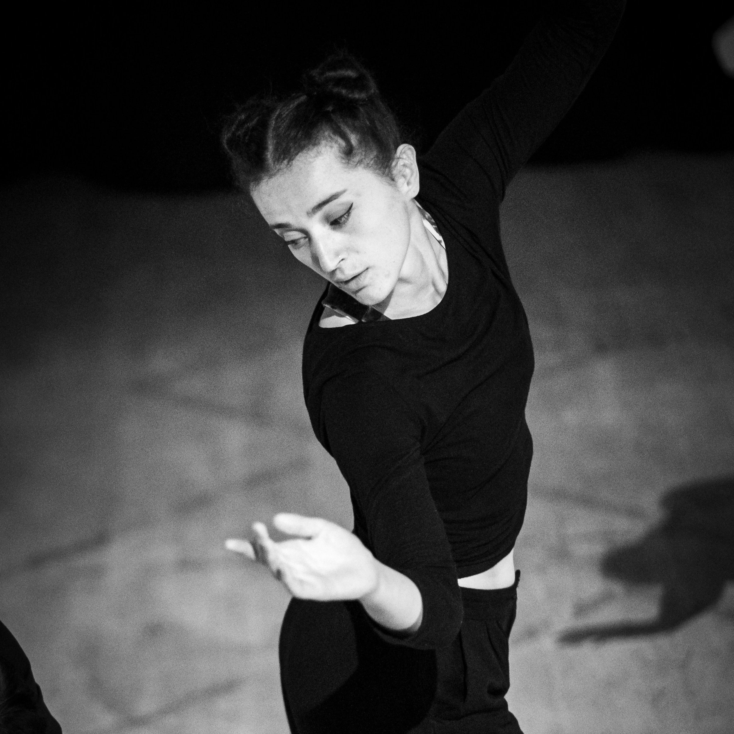  The image shows a female dancer in a studio, it looks like she is mid performance. The photo is black and white and almost is arial in it’s angle. She is wearing black clothing with dark hair tied up in two high buns. 