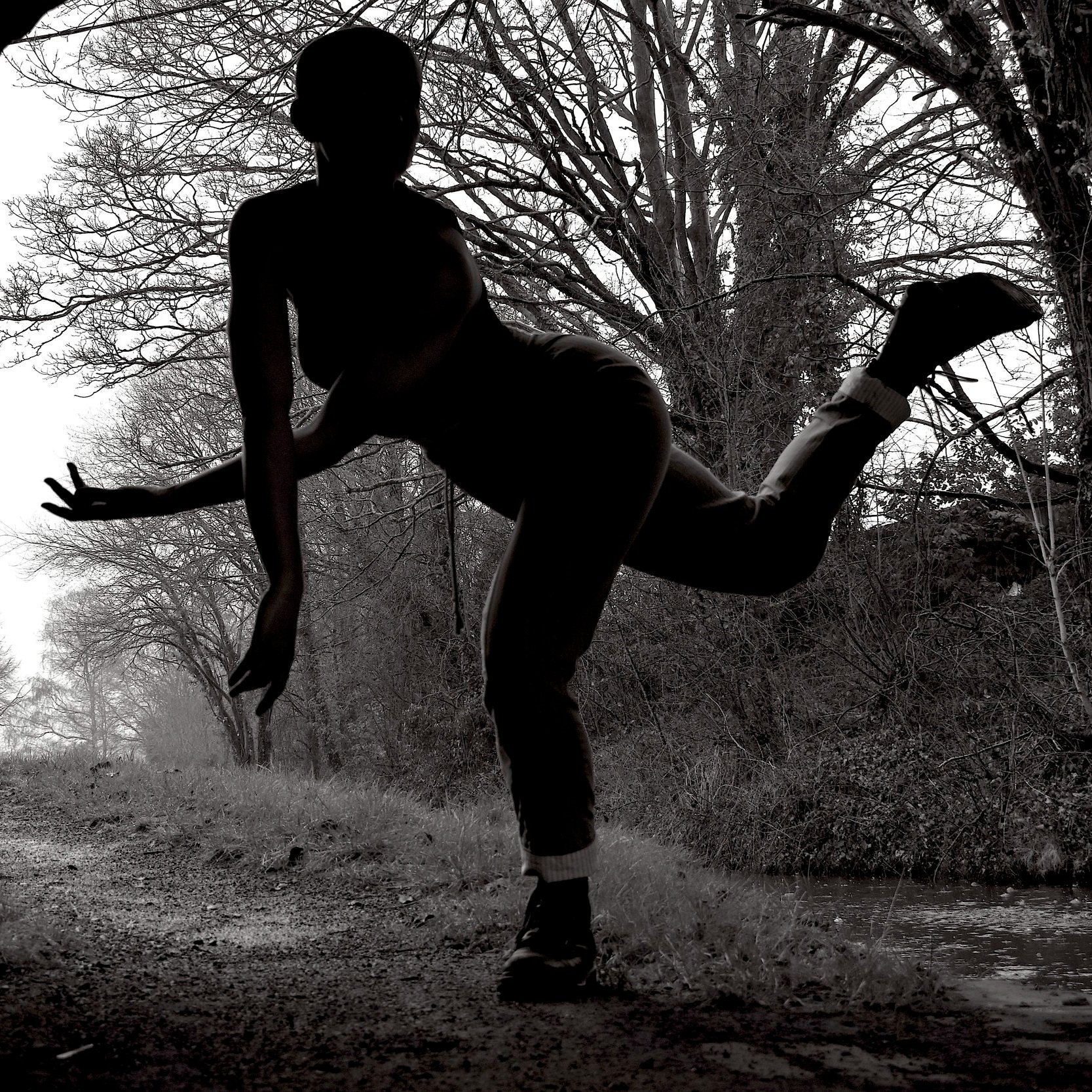  The image is black and white and shows a female dancer outdoors in a rural nature setting. She is mainly depicted as a silhouette. Balancing on leg she has the other leg up and behind her, it is bent. Her upper body is leaning forwards with arms low