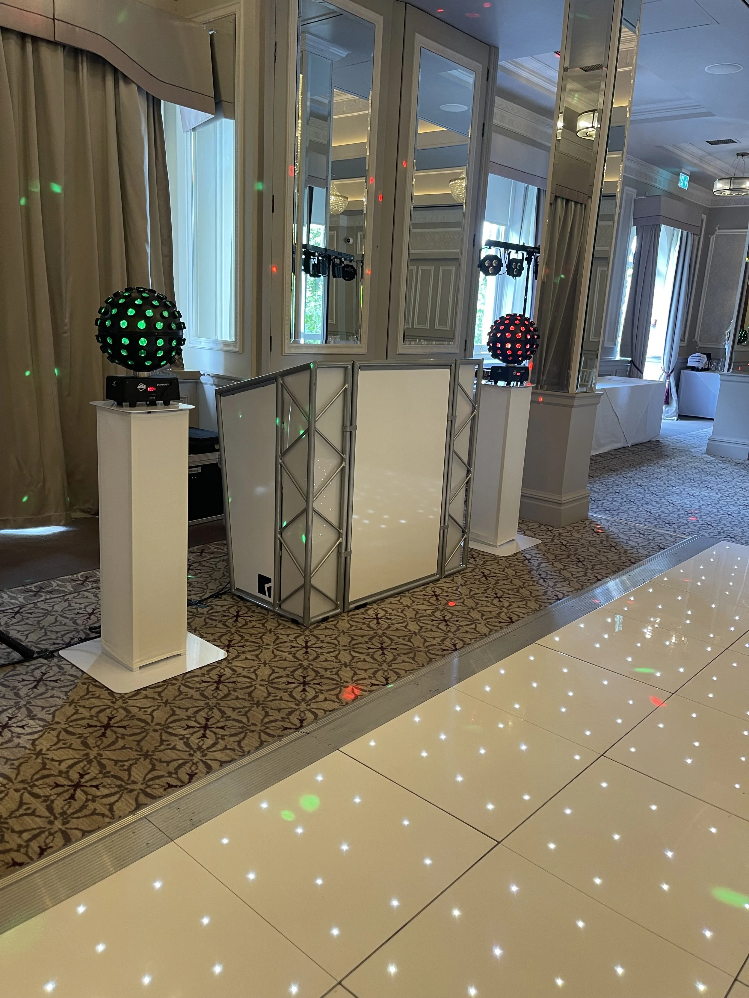 Party DJ setup with two colored disco light projectors on stands, a white DJ controller or mixer, and a white barrier or folding screen, in an elegant event hall with mirrored walls and patterned carpet.