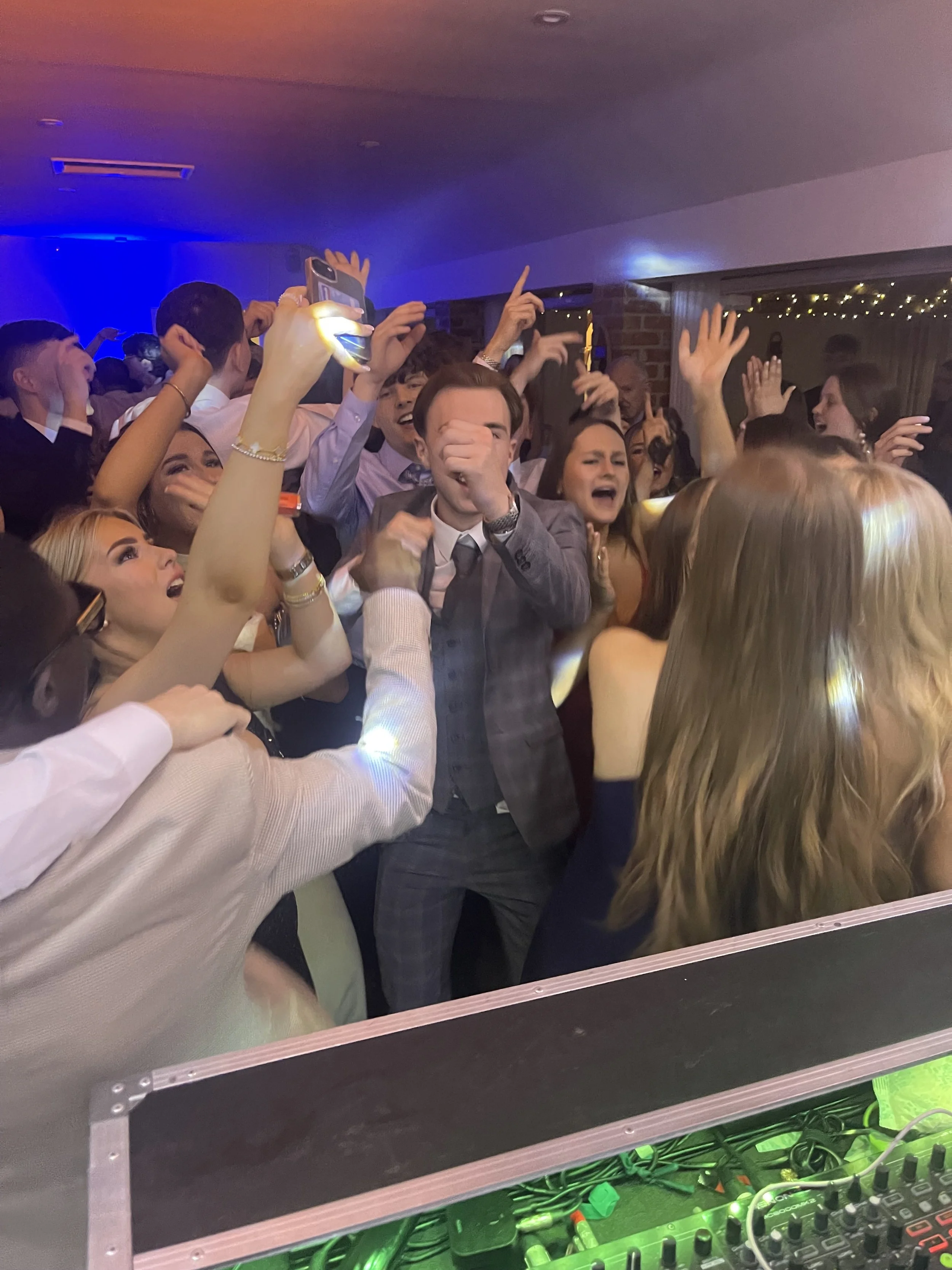 Group of young people dancing energetically at a party or event with colorful lighting and a DJ booth visible.