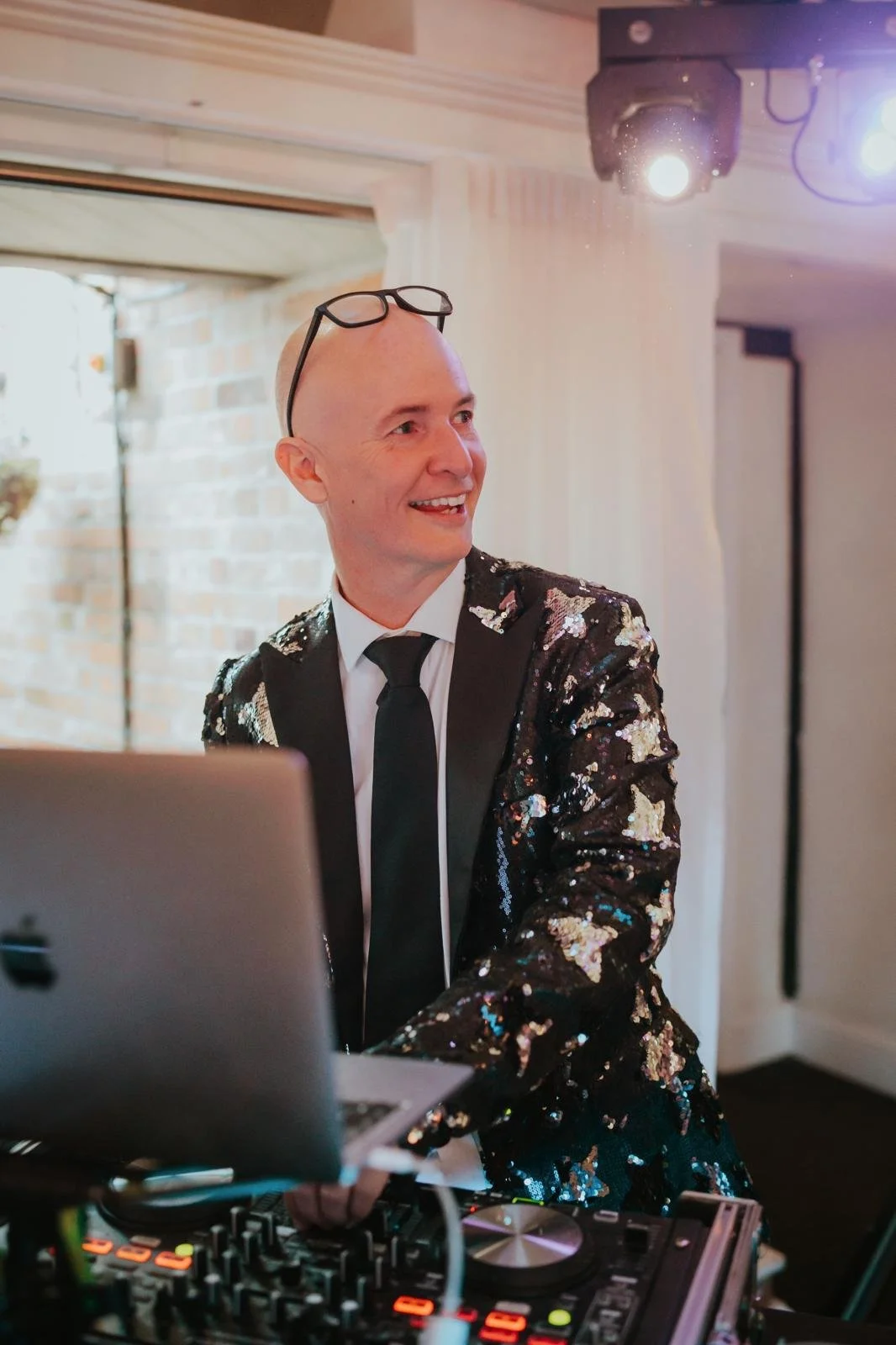 DJ wearing a sequined jacket, standing behind a DJ booth with a laptop and DJ equipment.