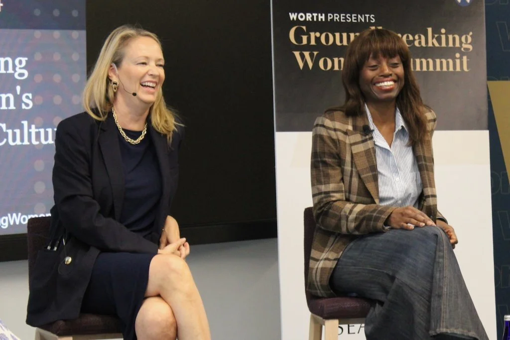 Two women are sitting on stage in front of a backdrop that reads "Groundbreaking Woman Summit." They are smiling and wearing business casual attire. One woman has blonde hair and is dressed in a black blazer and dress, while the other has dark hair and is wearing a plaid blazer and striped shirt.