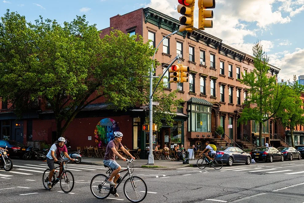 People riding bicycles across a Clinton Hill intersection with cars parked along the street.