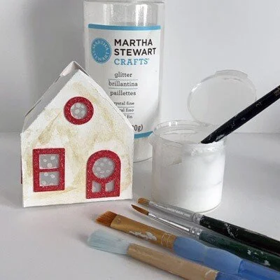 aged white cottage putz house with red window and door trim, paintbrushes, glue container and glitter