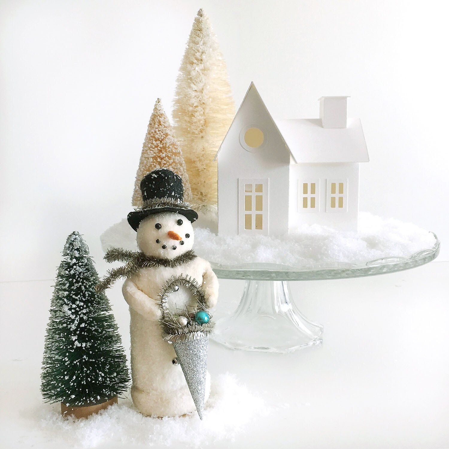 vintage felted snowman in front of a white full size farmhouse style putz house on a snow covered glass cake stand with two large white bottle brush trees.