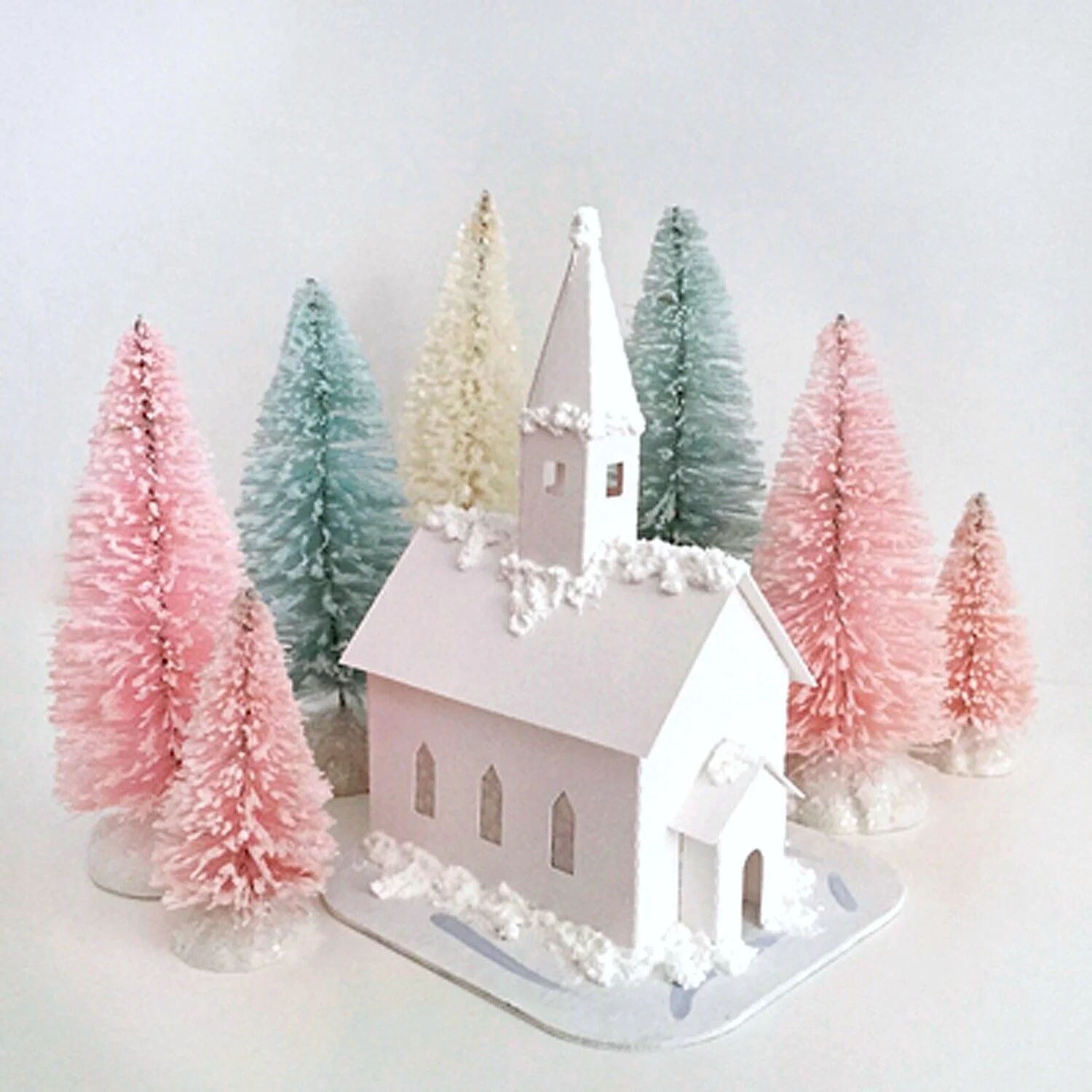 Miniature white church surrounded by pastel bottle brush Christmas trees in pink, blue, cream, and green, arranged on a white surface.