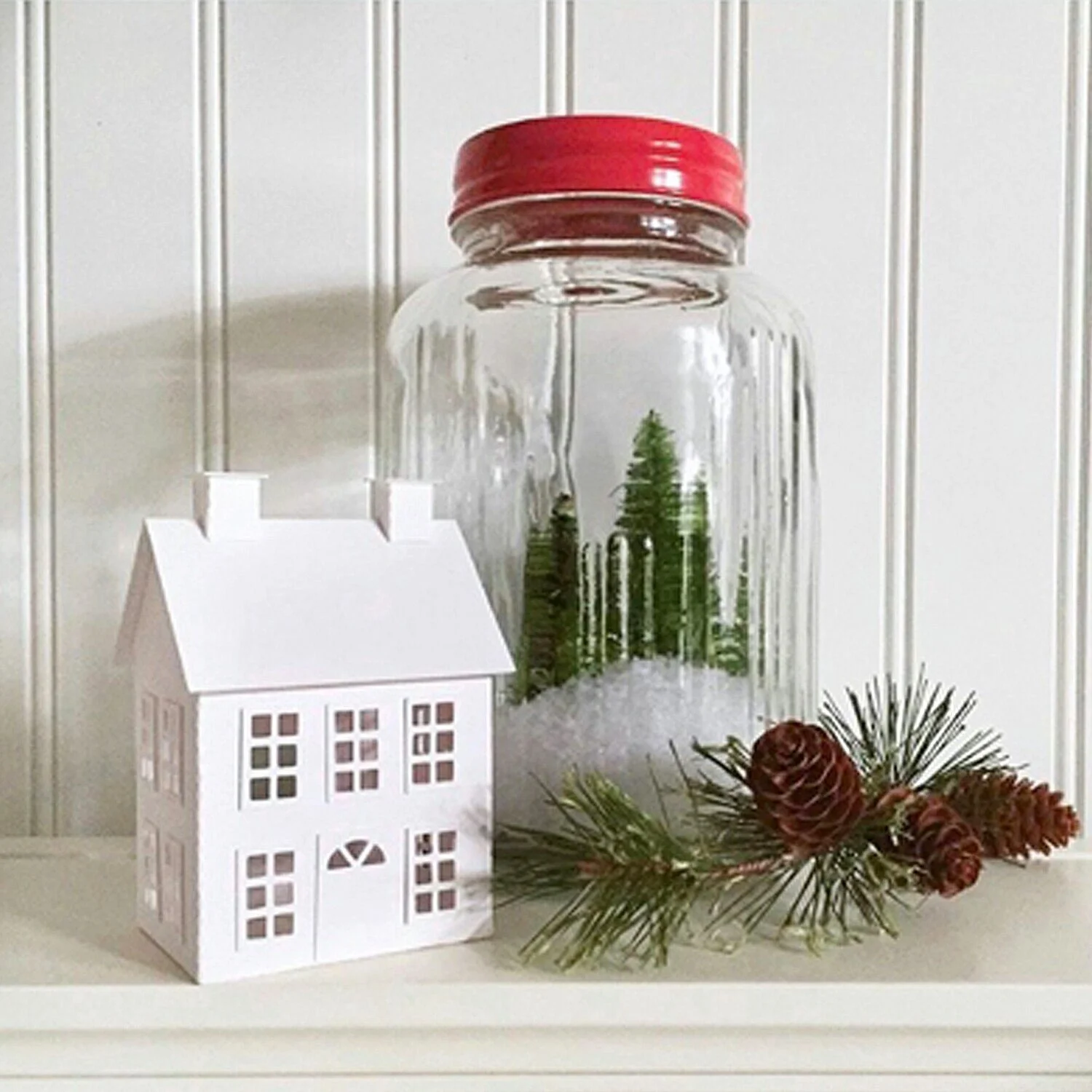 Decorative Christmas scene of a white prim style paper house next to a glass jar filled with snow and small bottle brush trees and a pine branch with pinecones, on a white shelf against a white bead board wall.