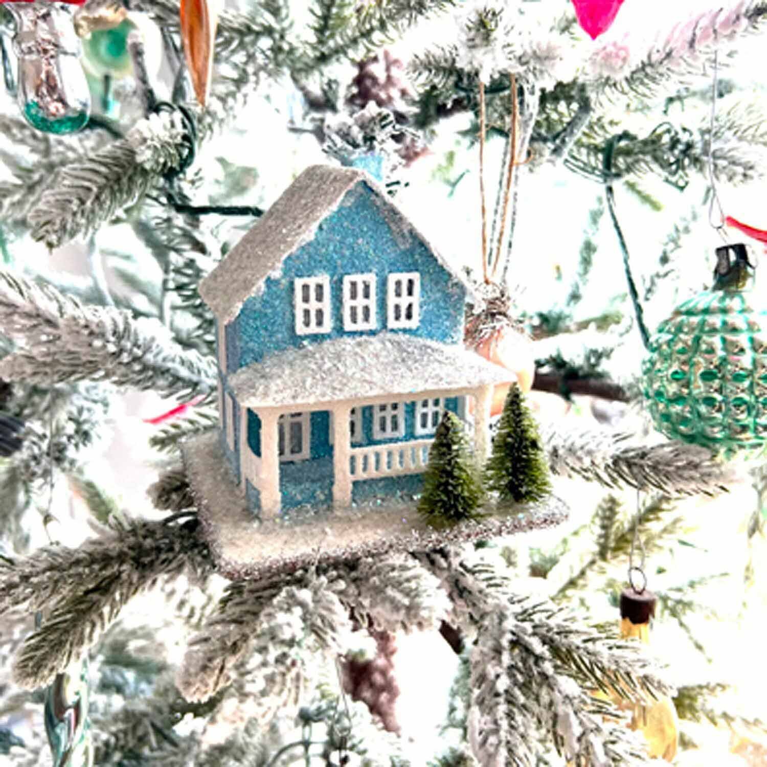 Glittered blue putz house ornament on a Christmas tree among other glass ornaments.