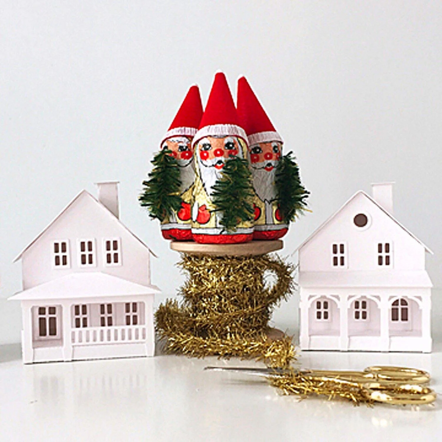 Christmas elf figures with red hats and white beards on a wooden spool of gold tinsel garland sit between two white paper houses with front porches. 