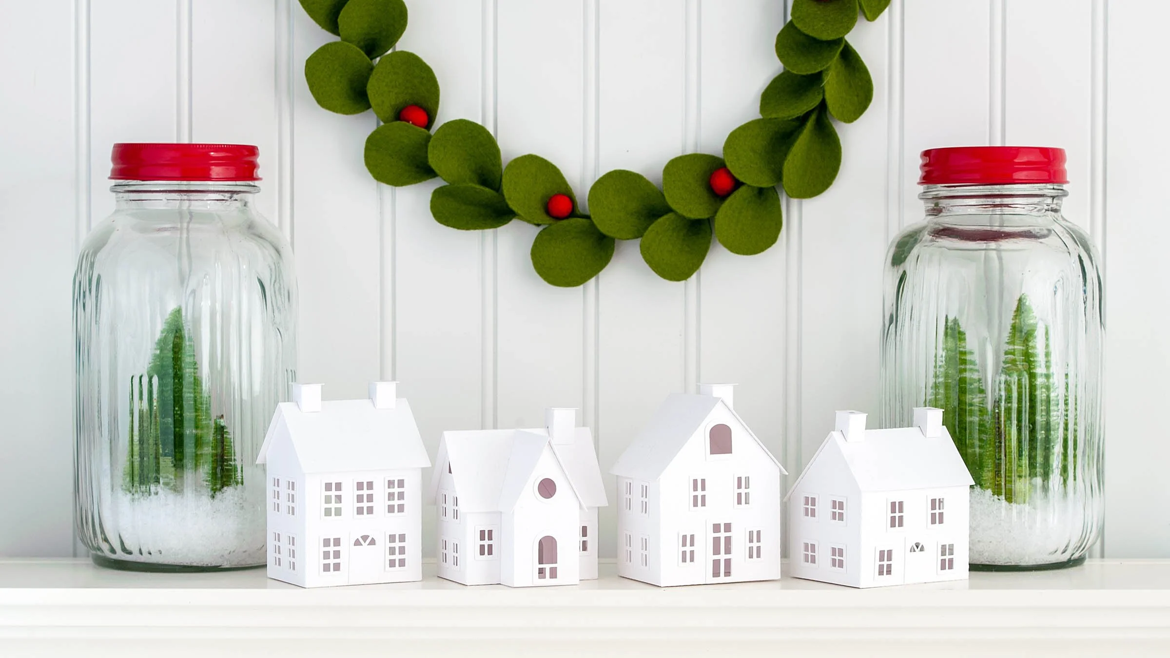 four white paper houses between 2 glass jars filled with snow and bottle brush trees on a white mantel in front of a white beadboard wall with a green felt wreath hanging above.