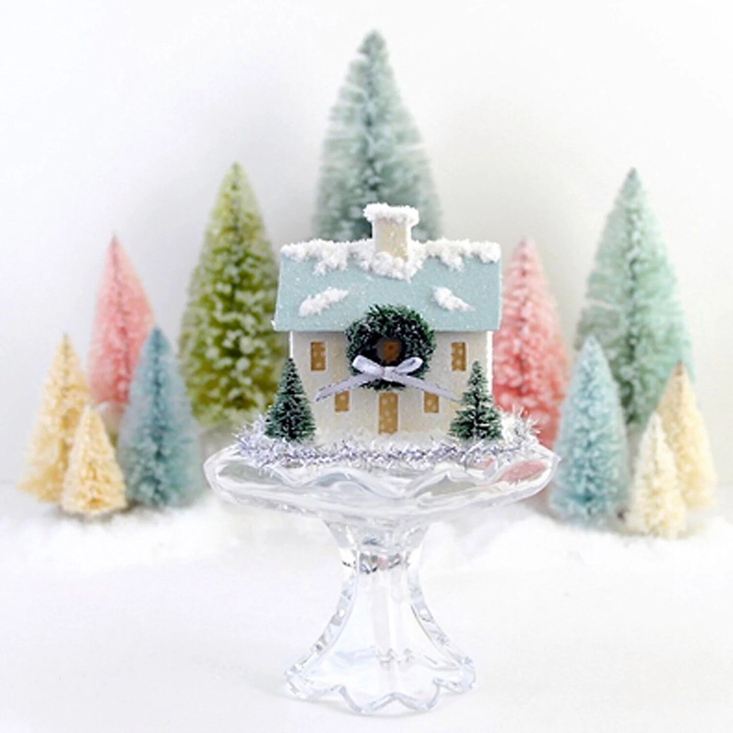 Miniature snowy Christmas house with a teal blue roof, green wreath and bow on a glass cake stand, surrounded by pastel bottle brush trees and snow.
