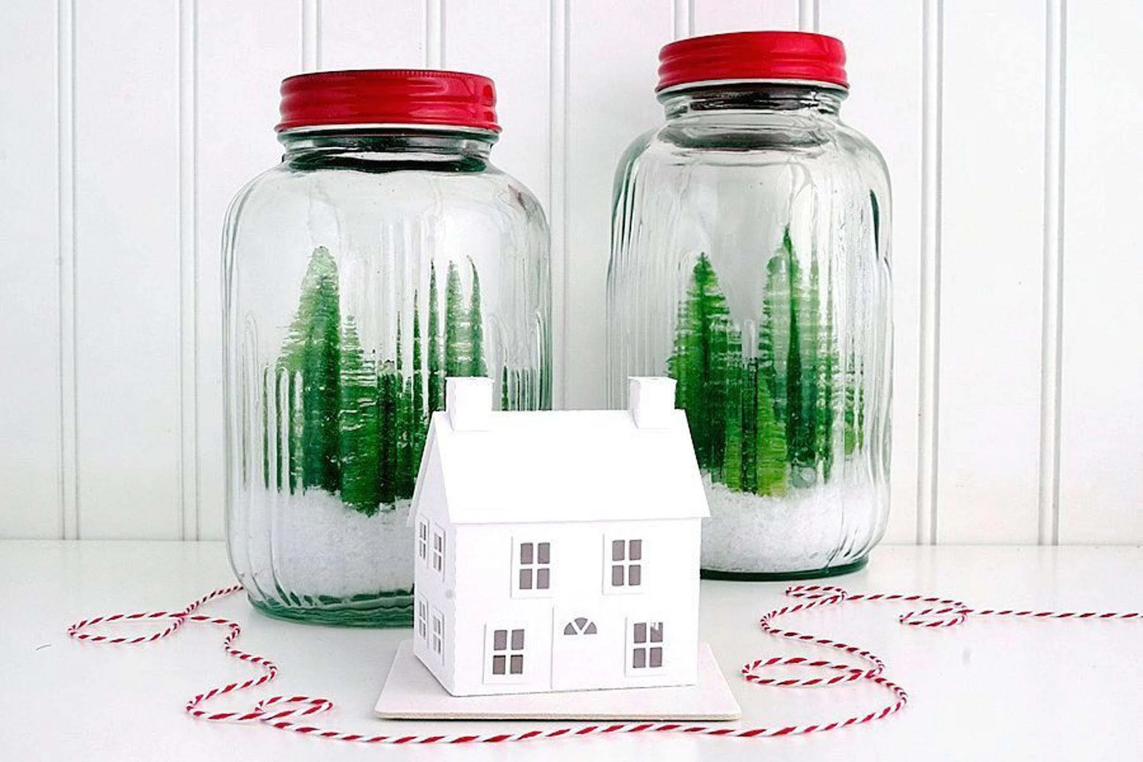white new england salt box putz house in front of two ribbed glass jars filled with snow and bottle brush trees
