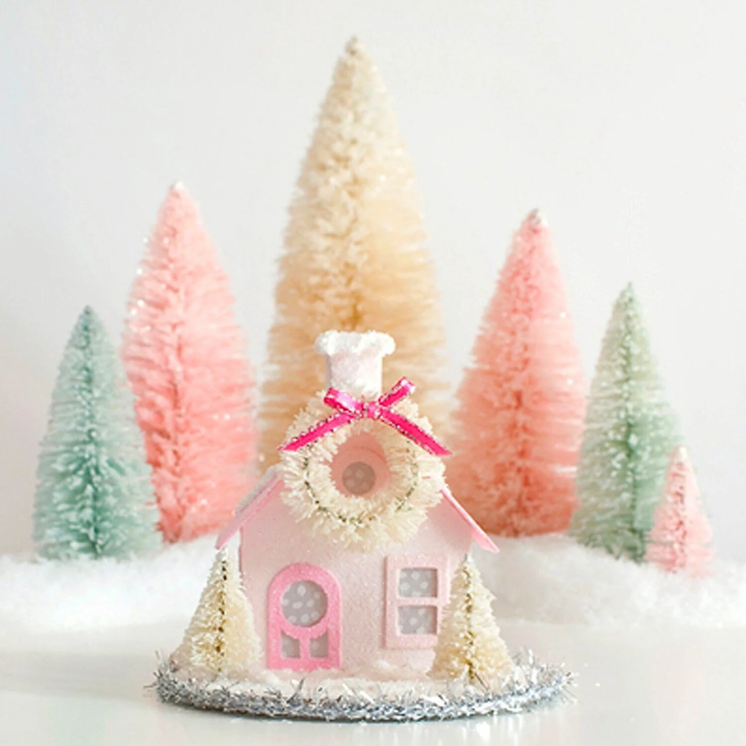 Glittered miniature pink and white putz house with a wreath and pink bow, surrounded by pastel-colored bottle brush trees, set on a snowy surface.