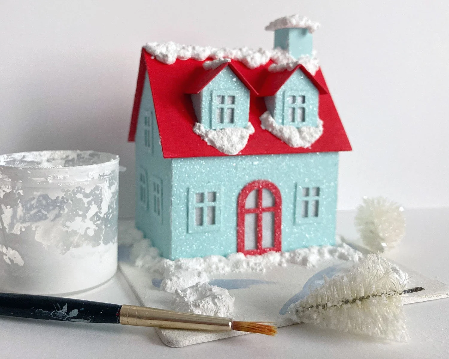 turquoise putz house with a red door next to a container of textured snow and a paintbrush