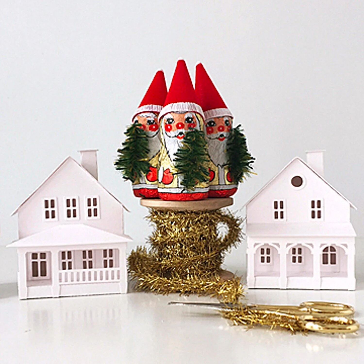 Three Christmas elf decorations wearing red Santa hats with faces painted with red noses and rosy cheeks, standing on a round wood spool of gold tinsel, with white paper houses on either side.