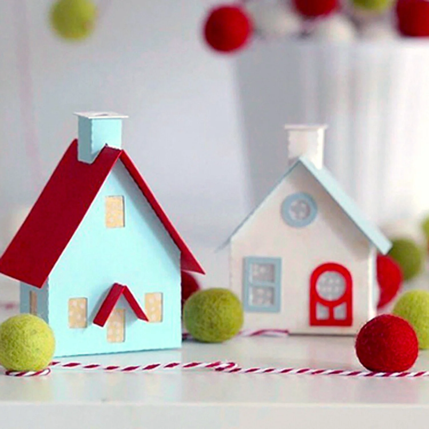 Colorful miniature putz houses with red and blue roofs, surrounded by a bakers twine garland of green and red felt balls on a white surface.