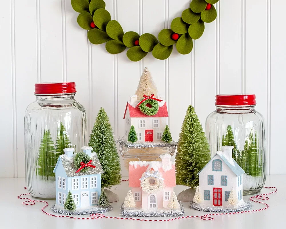 Miniature Christmas village decorated houses, bottle brush trees, and glass jar decorations, set against a white beadboard wall with a green felt wreath with red berries hanging above.