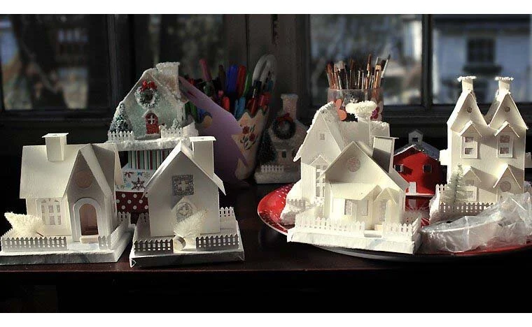 five white putz houses on bases with picket fences sitting on a table waiting to be glittered