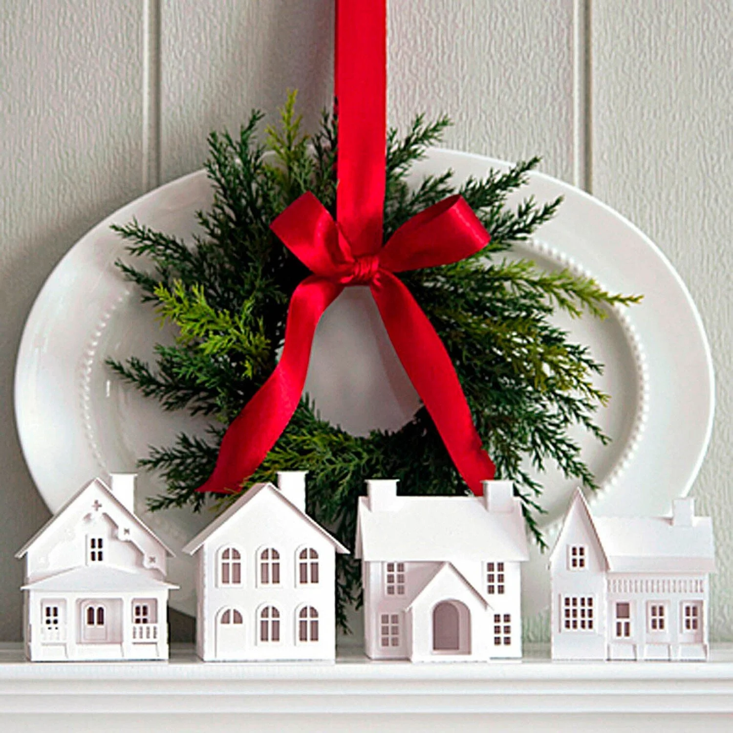 White miniature paper houses in front of a white ironstone platter on a white fireplace mantel, with a Christmas wreath made of greenery and a red bow hanging above them.
