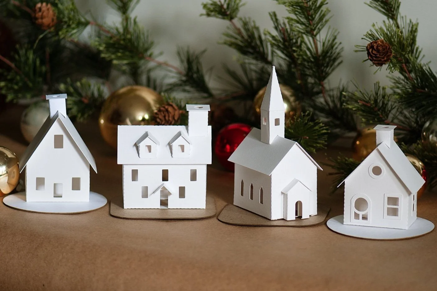 close up of white christmas village houses in front of greenery and glass ornaments