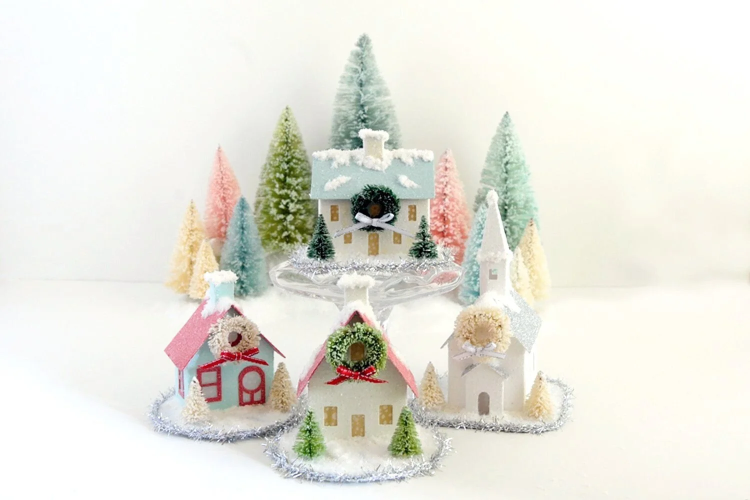 three decorated christmas village house kits and church with snow and multicolored bottle brush trees,on a white background.