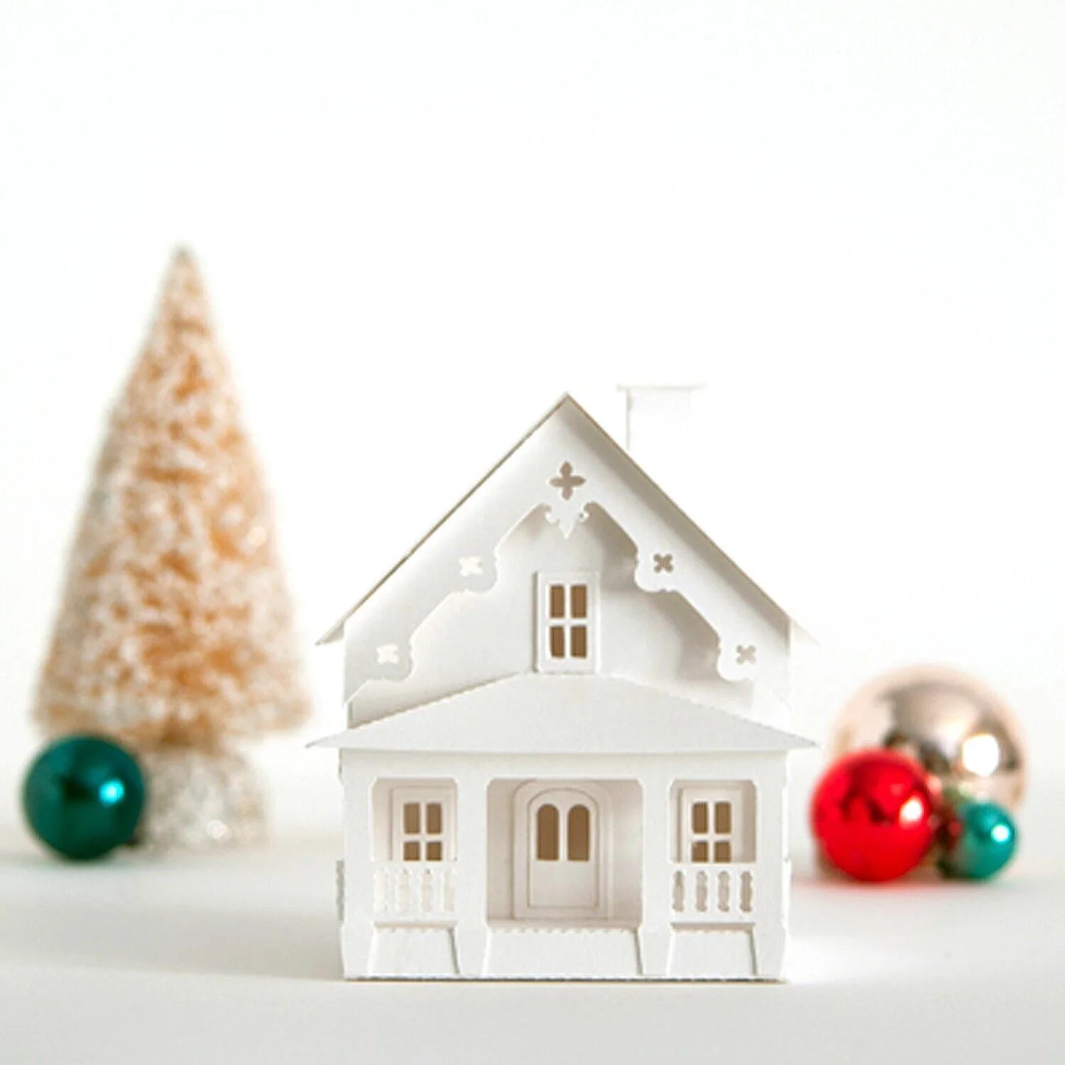 Miniature white putz house kit  with gingerbread trim and front porch between a small snow-covered tree and red, silver and teal vintage glass ornaments against a white background.