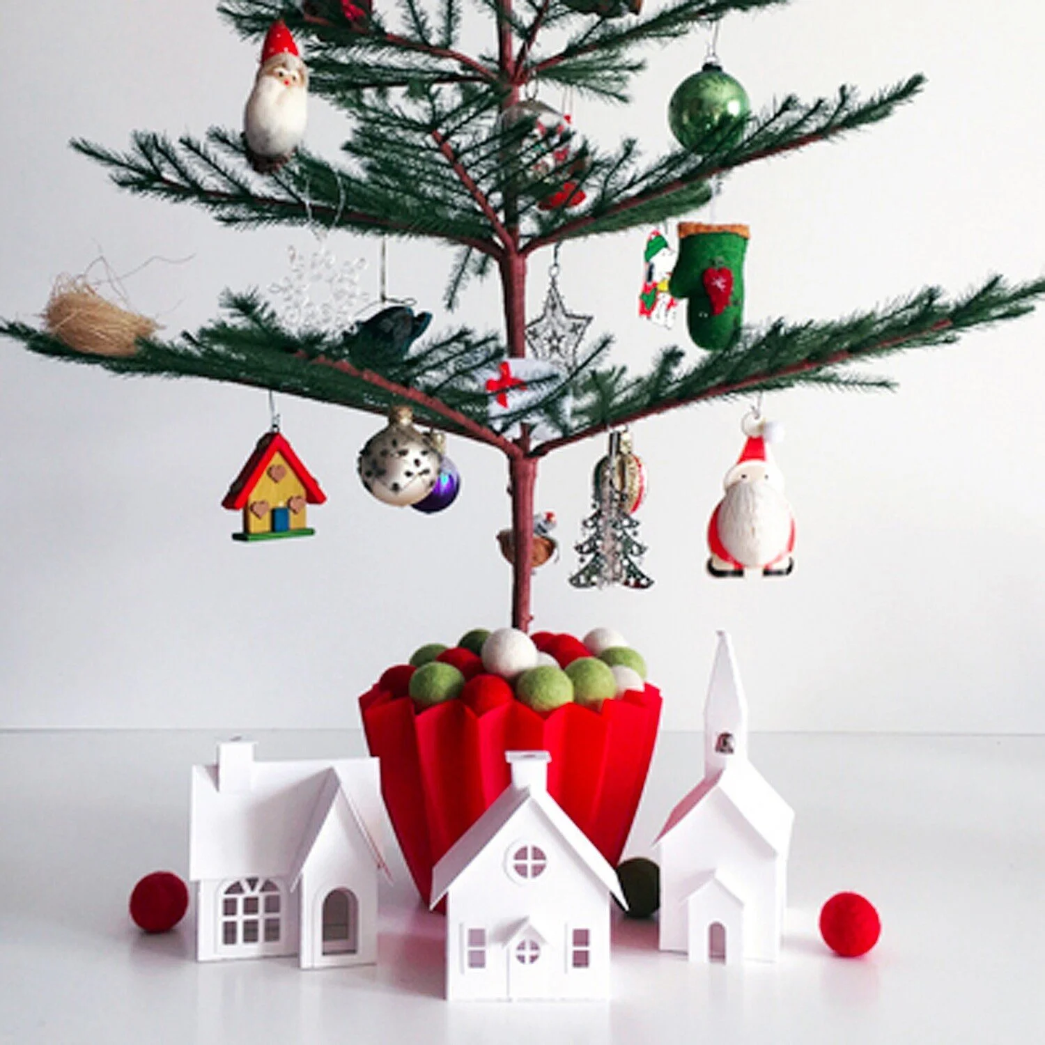 Christmas Feather Tree decorated with small handmade and glass ornaments in a red container surrounded by three white houses and felt balls.