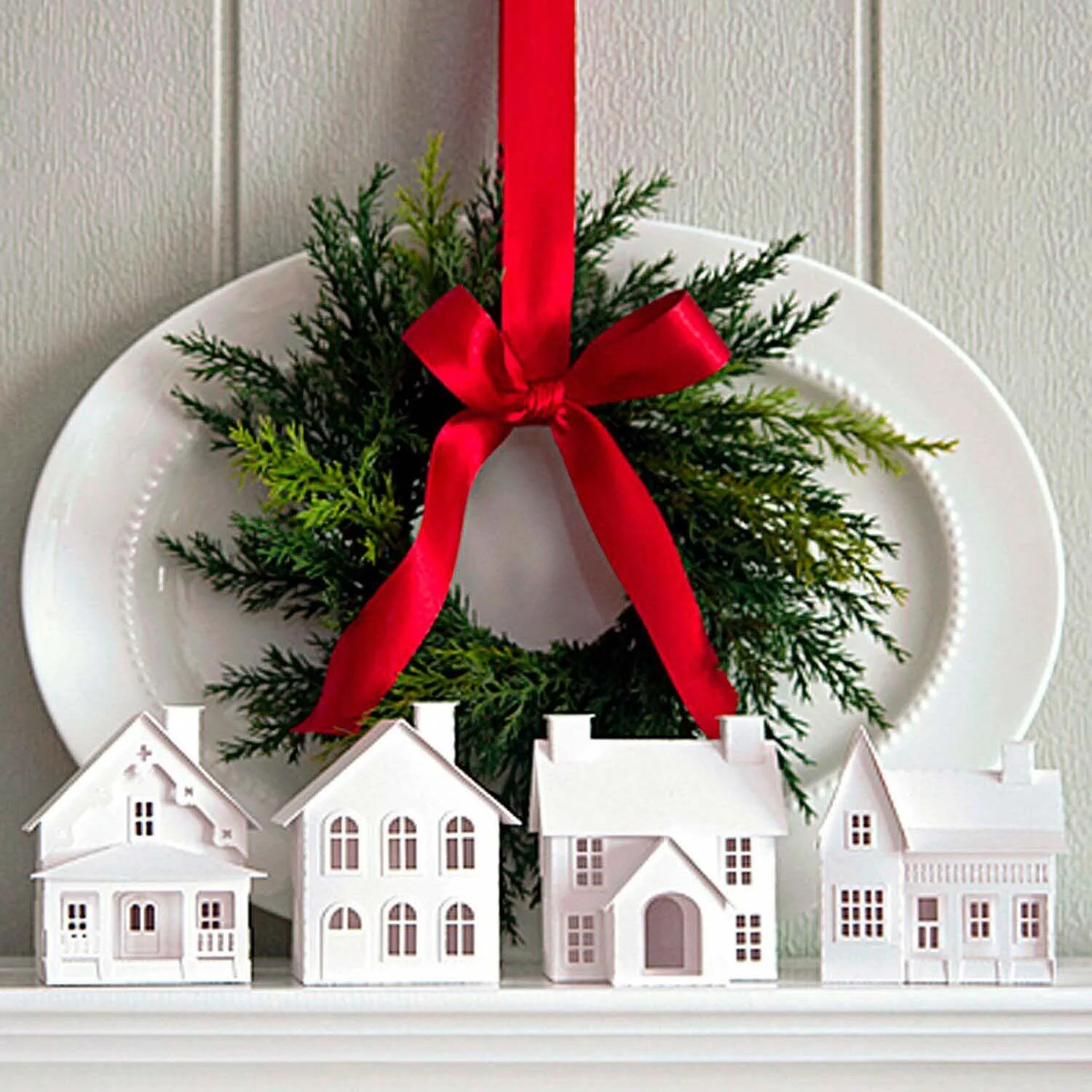 A white wall featuring a decorative Christmas wreath with greenery and a red ribbon, hung above a row of small white ceramic houses with cut-out windows and doors.