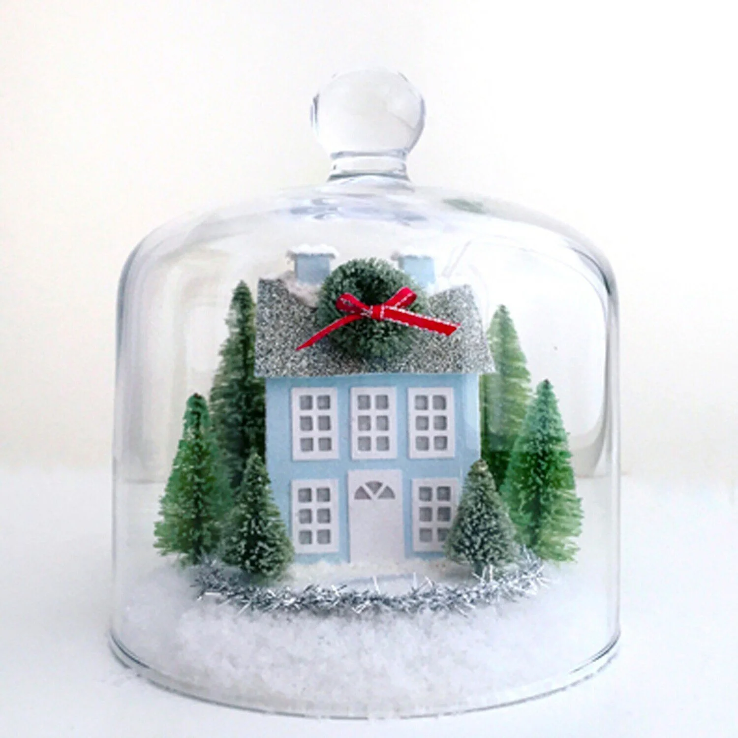 Blue Prim Christmas village house with a small green wreath and red bow on the chimney surrounded by green bottle brush trees inside a glass cloche.