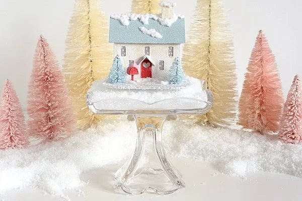 Valentine Colonial house with teal blue roof, red door with a heart window and mailbox and 2 turquoise bottle brush trees sitting on a small cake pedestal in front of white and pink bottle brush trees.
