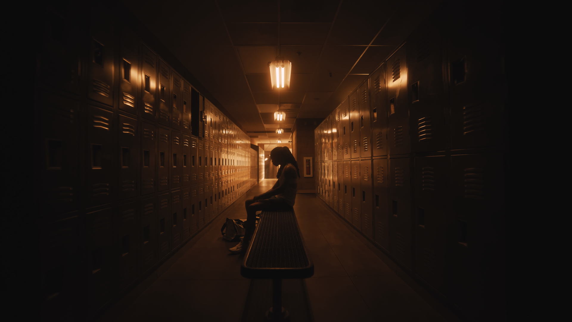 Silhouette of a girl sitting alone on a bench in a school locker hallway with lockers on both sides, illuminated by fluorescent lights overhead.