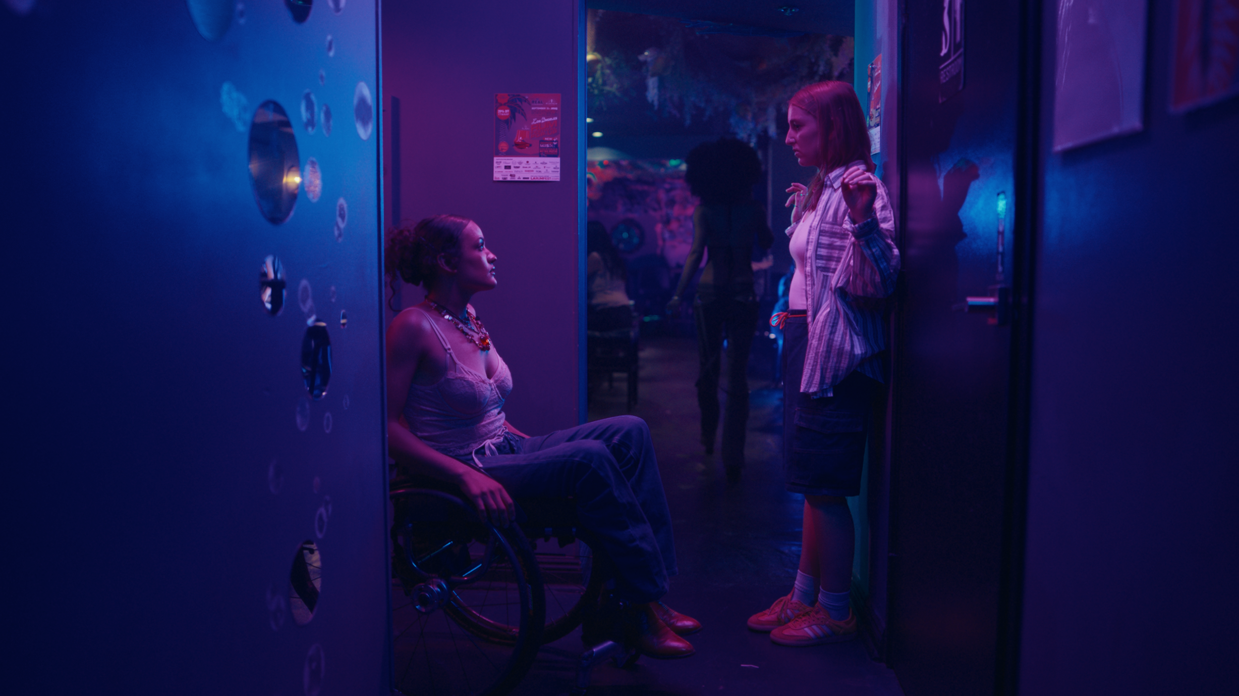 Two young women are in conversation in a dark, neon-lit environment, one sitting in a wheelchair and the other standing, near a row of lockers with circular vents.