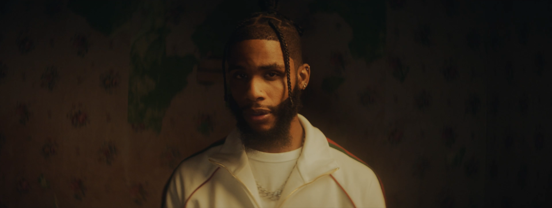 A man with braided hair and a beard, wearing a white jacket with red and green stripes, looking at the camera in a dimly lit setting.